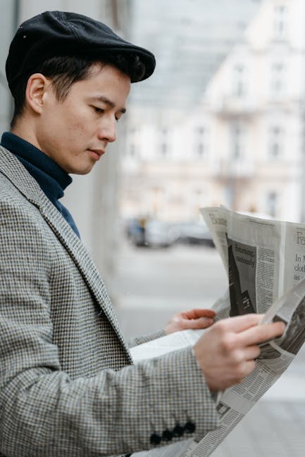 Young man wearing a blazer and flat cap, reading a newspaper on the street with blurred buildings in the background.