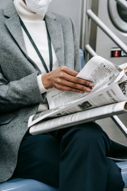 Crop anonymous black female entrepreneur in elegant outfit and medical mask reading newspaper while riding train