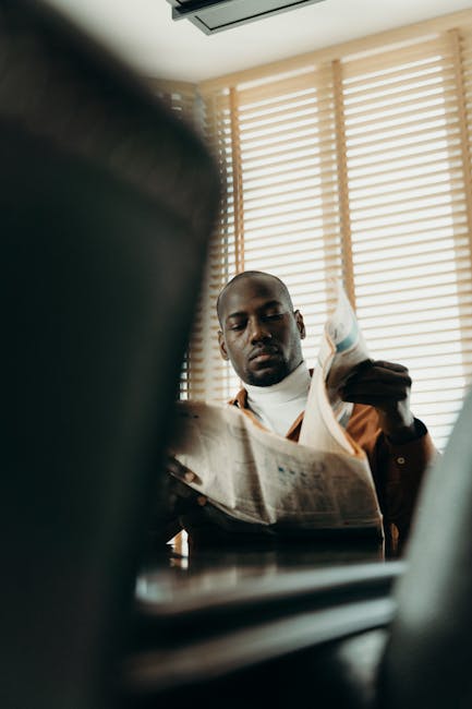 A man reads a newspaper sitting at a table inside with window blinds.