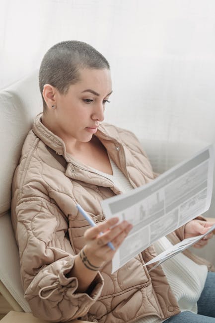 Young bald woman attentively reading newspaper indoors, holding pen for note-taking.